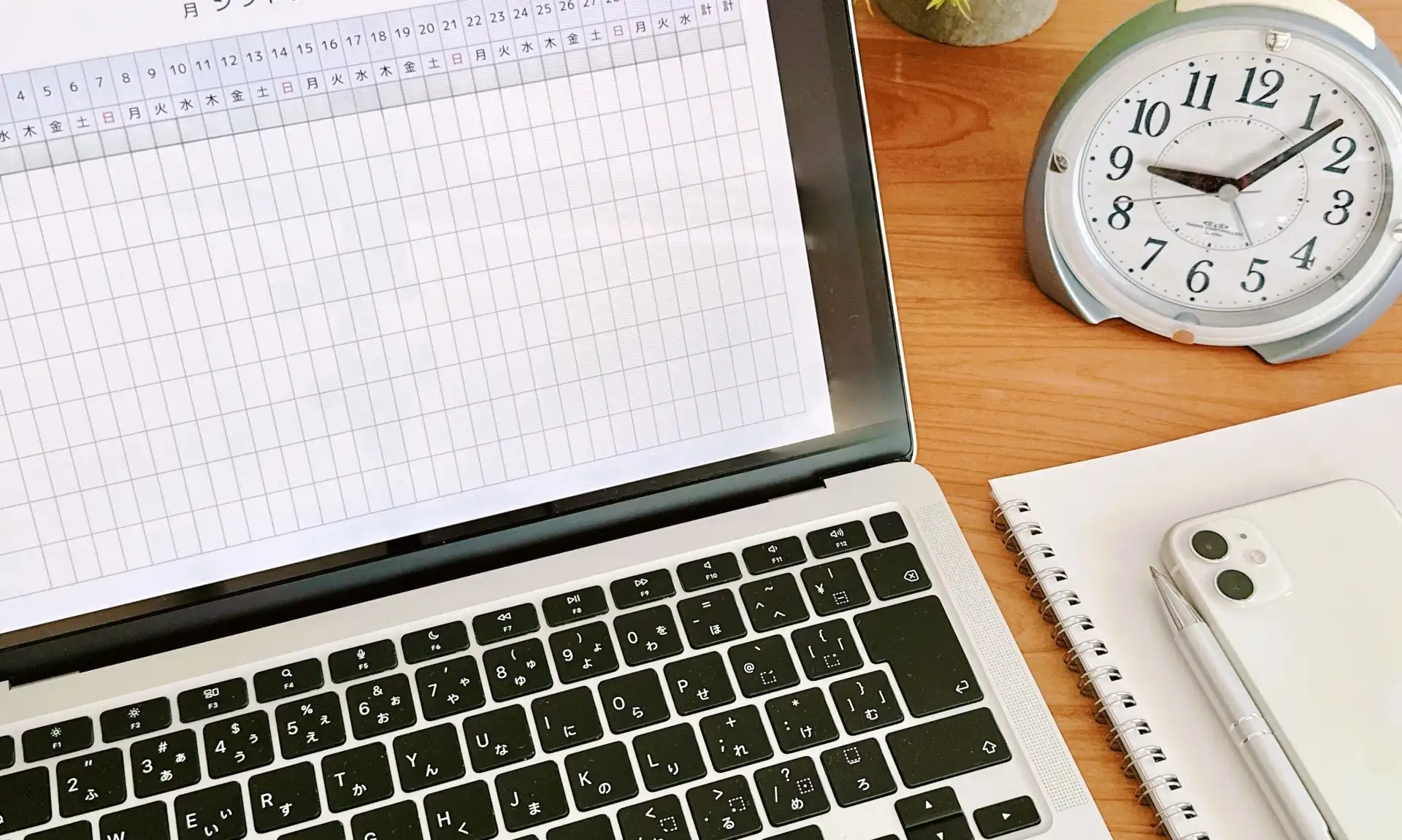 エクセル表を開いているPCの画面と目覚まし時計、文房具